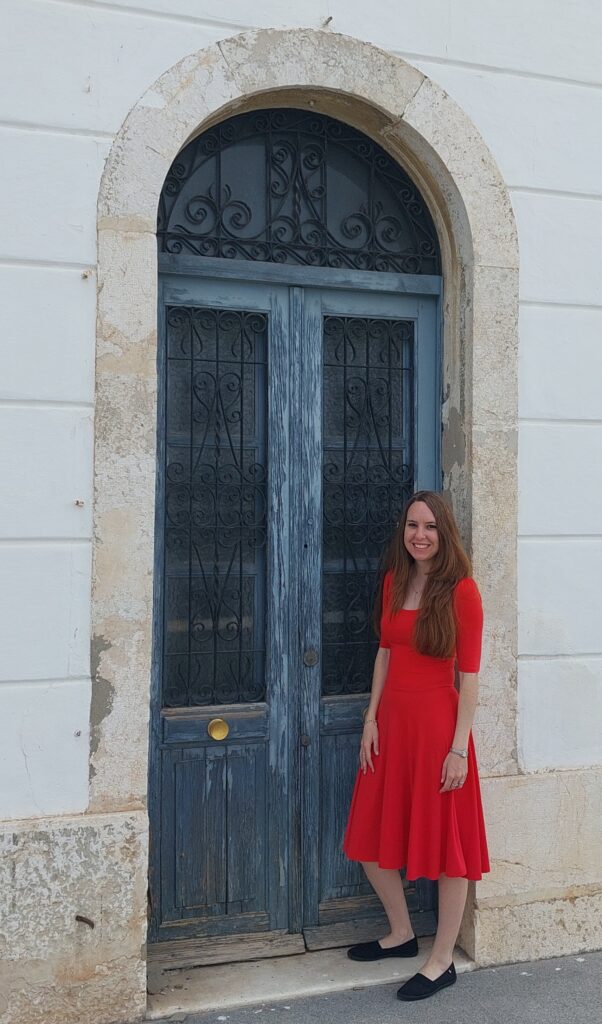 Elizabeth Olmedo wearing a coral dress stands in front of an old, smokey-blue door.